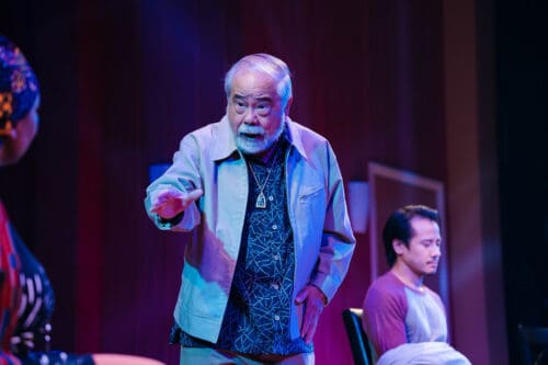 Older man with white beard speaking and pointing on stage, younger man seated nearby.