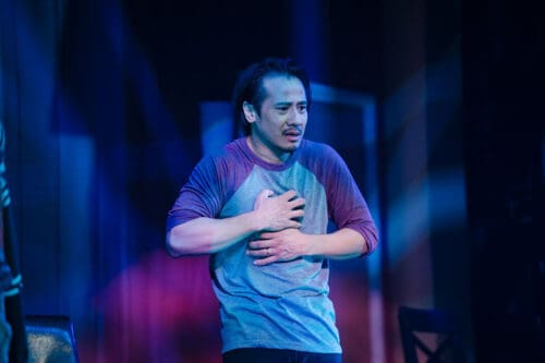 Man clutching his chest in distress on stage under blue and purple lights.