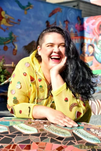 A joyful woman with dark hair wearing a yellow sweater with a smiley face pattern.