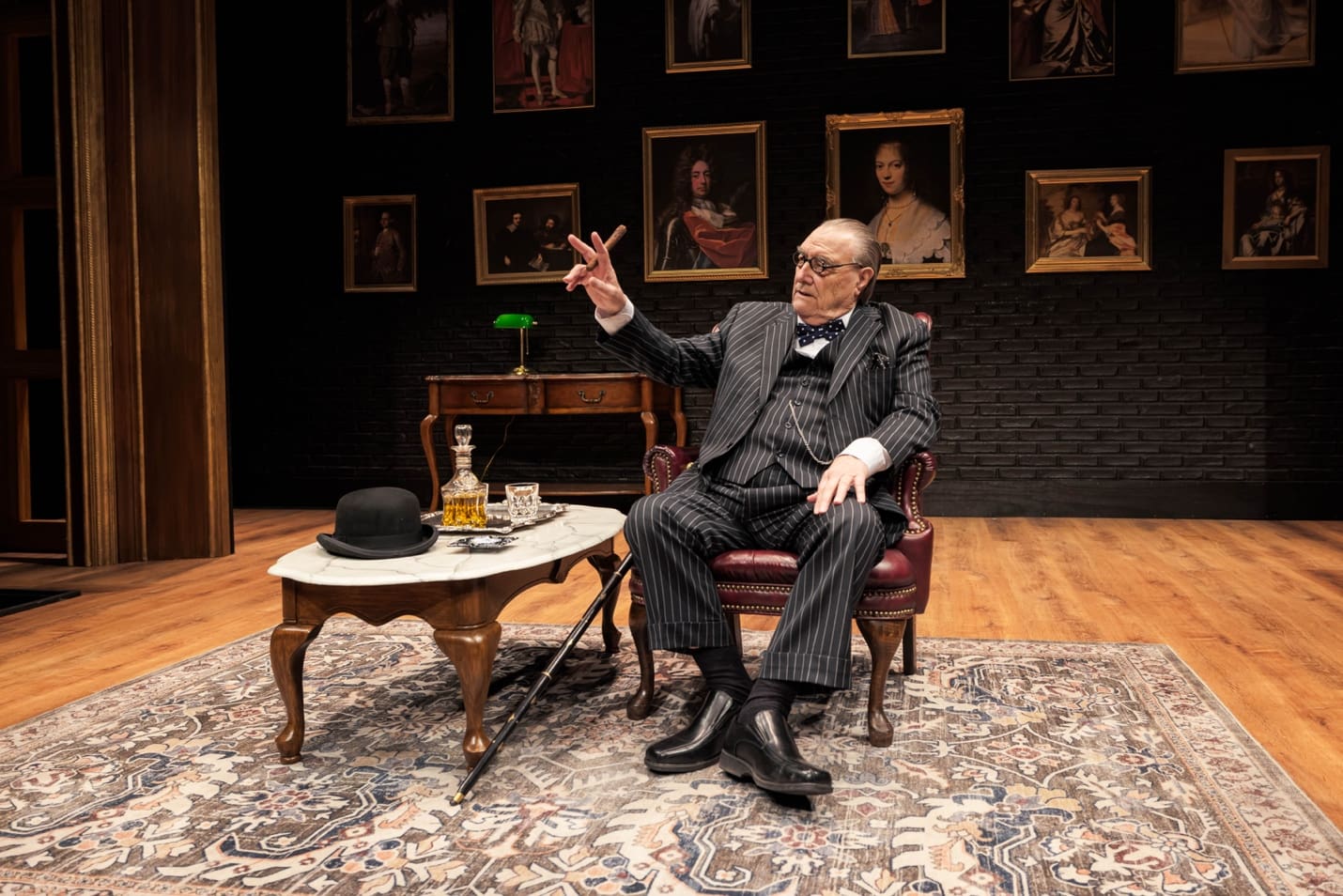An elderly man gestures while seated in a classic study setting.