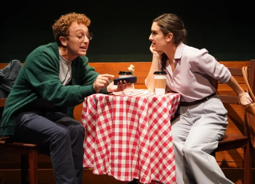 Two actors intensely converse over a checkered tablecloth on stage.