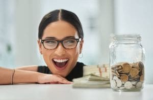 Excited woman with glasses smiling near a jar of coins and cash.