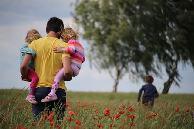 A man holding a child with another child running in a field of flowers.