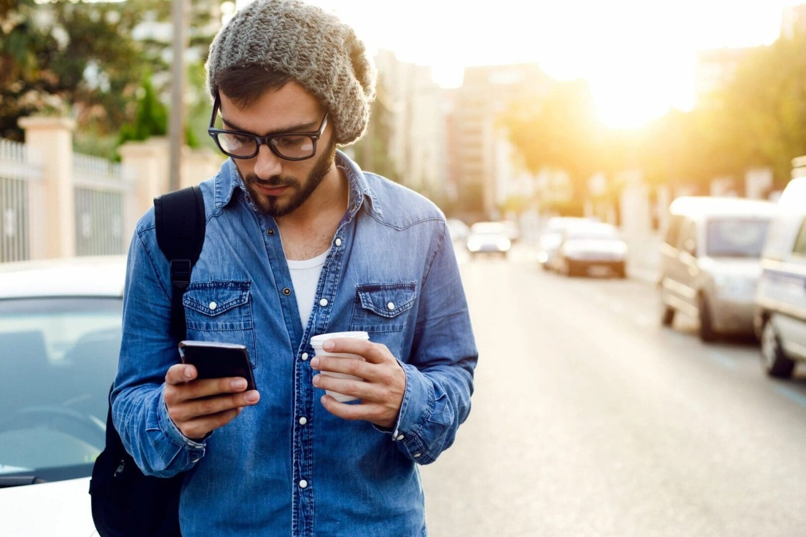 Young man with glasses and a beanie checks his phone while holding a coffee.