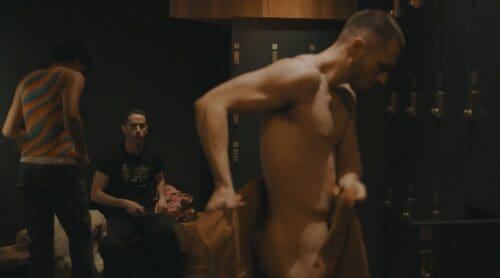 A shirtless man adjusting his pants in a dimly lit room with two seated people watching.