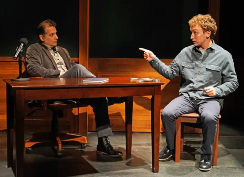 Two men engaged in a serious conversation at a wooden table in a dimly lit room.
