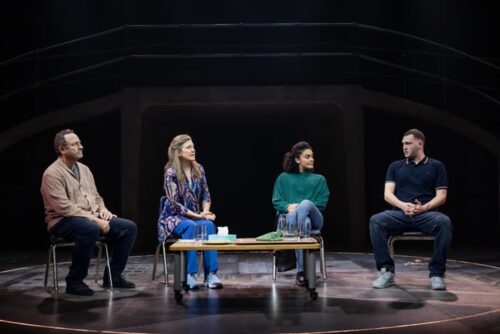 Four actors seated on stage engaged in a serious conversation during a theatrical performance.