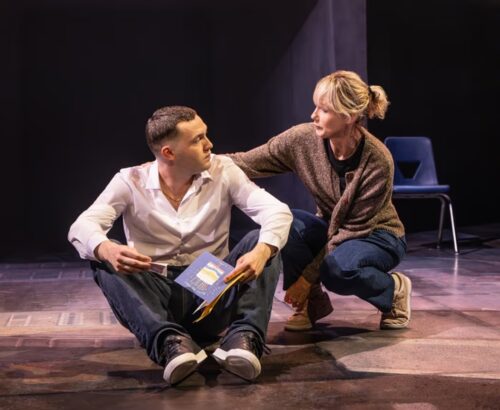 A man sits on stage holding a book while a woman kneels beside him, offering comfort.