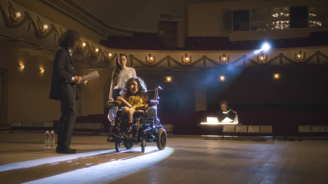 Actress with a disability driving around the stage in an electric wheelchair and rehearsing a performance with actors and a director in a dim theater, with a spotlight.