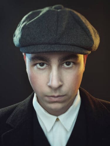 Close up portrait of real man in black suit and flat cap against black background. Caucasian male model in peaky blinders style staring at camera. Photography from a DSLR camera. Sharp focus on eyes.